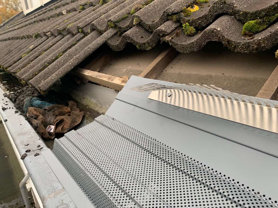Dakgoot met geïnstalleerde dakgootroosters ter voorkoming van verstopping door bladeren en als vogelwering, naast een dak met dakpannen waarop mos groeit.
